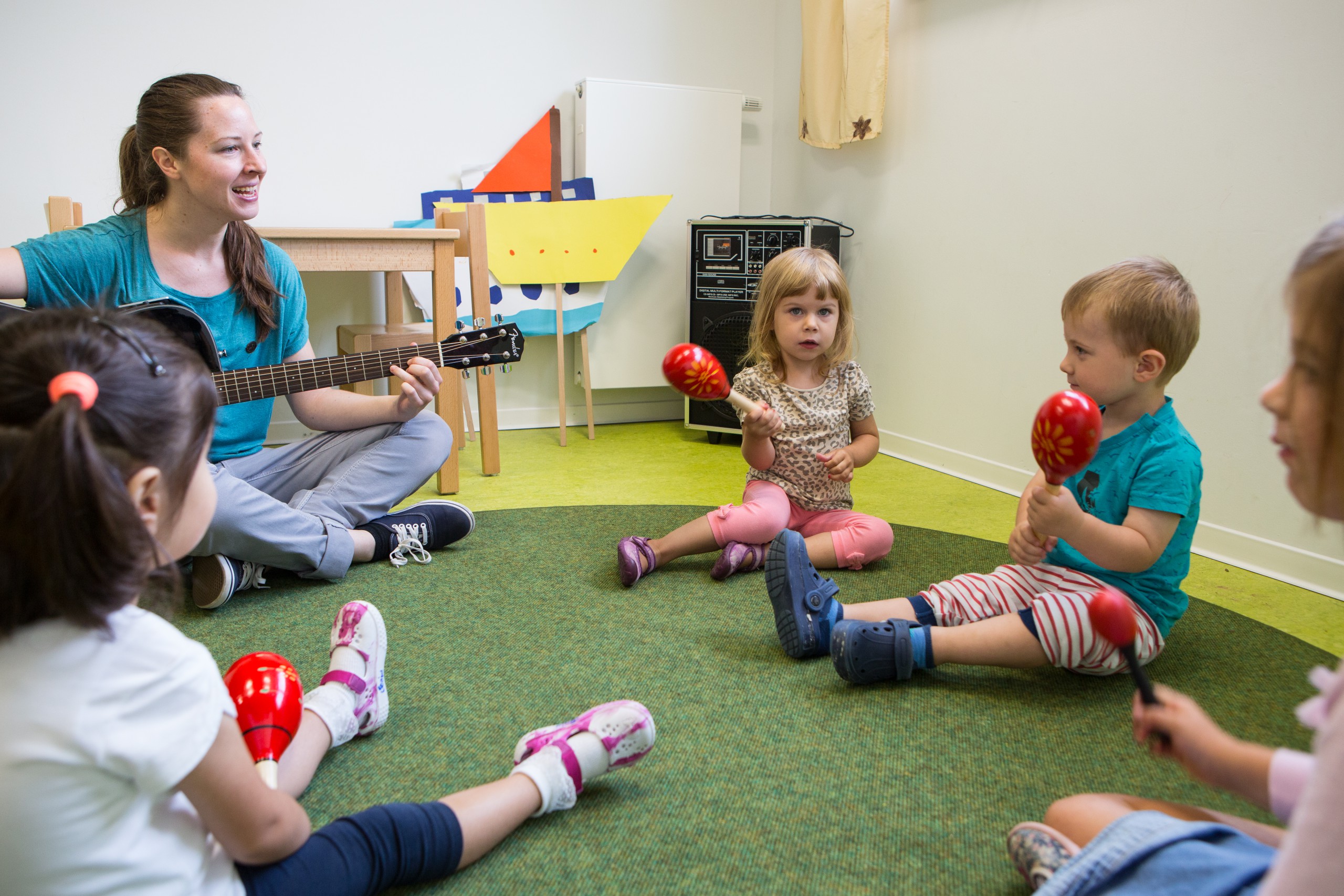 Kinder musizieren im Kreis mit Rasseln, eine erwachsene Person spielt Gitarre