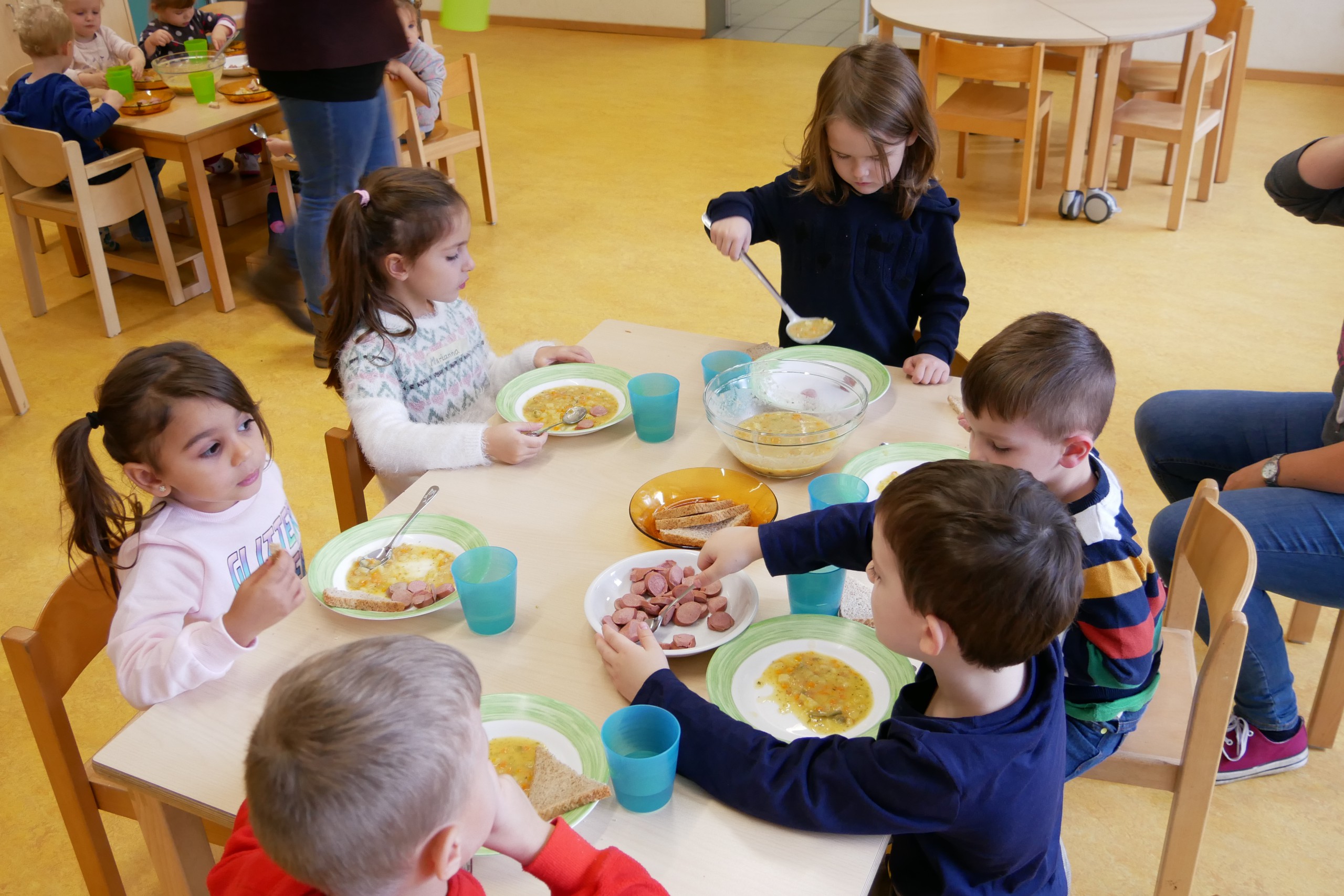 Kinder sitzen beim Mittagessen um einen Tisch und schöpfen sich selbständig das Essen auf den Teller