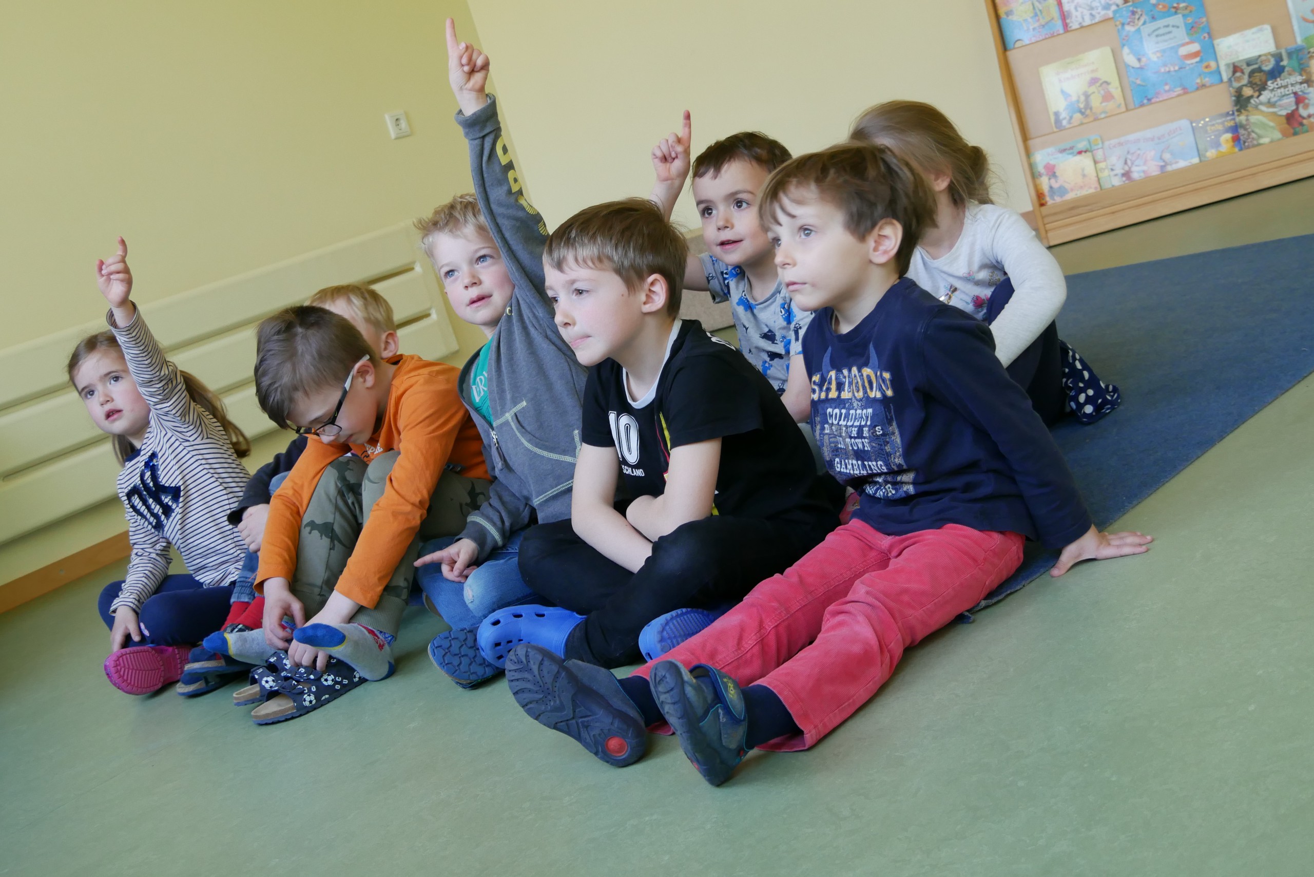 Mehrere Kinder sitzen auf dem Boden und schauen in eine Richtung. Zwei von ihnen heben die Hand, um sich zu melden