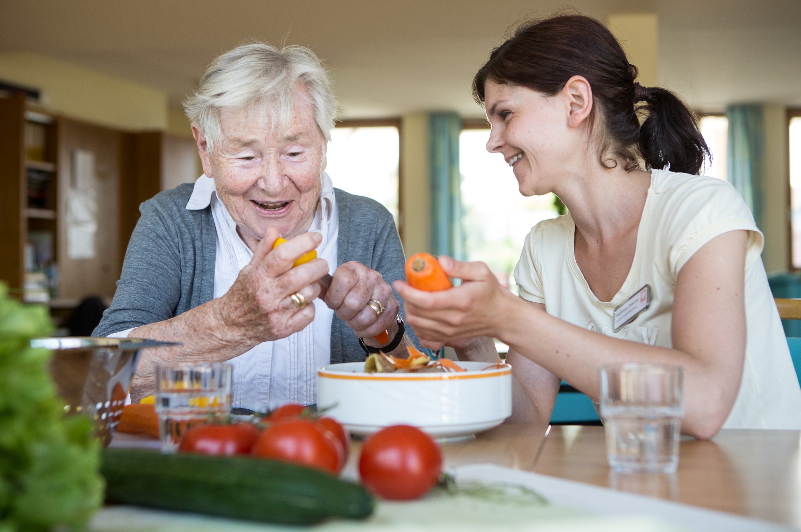 Eine ältere und eine jüngere Person sitzen lachend an einem Tisch und schälen Gemüse